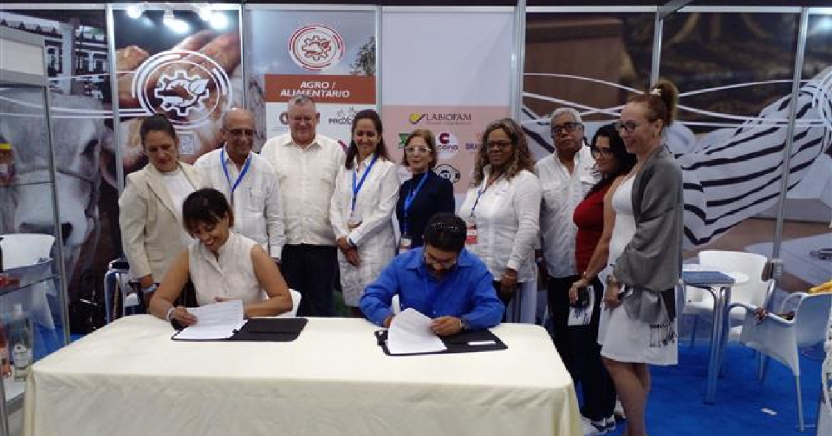 Magaly Iserne, de pie con chaqueta azul, y Jorge Guerra, firmando el convenio. © Prensa Latina