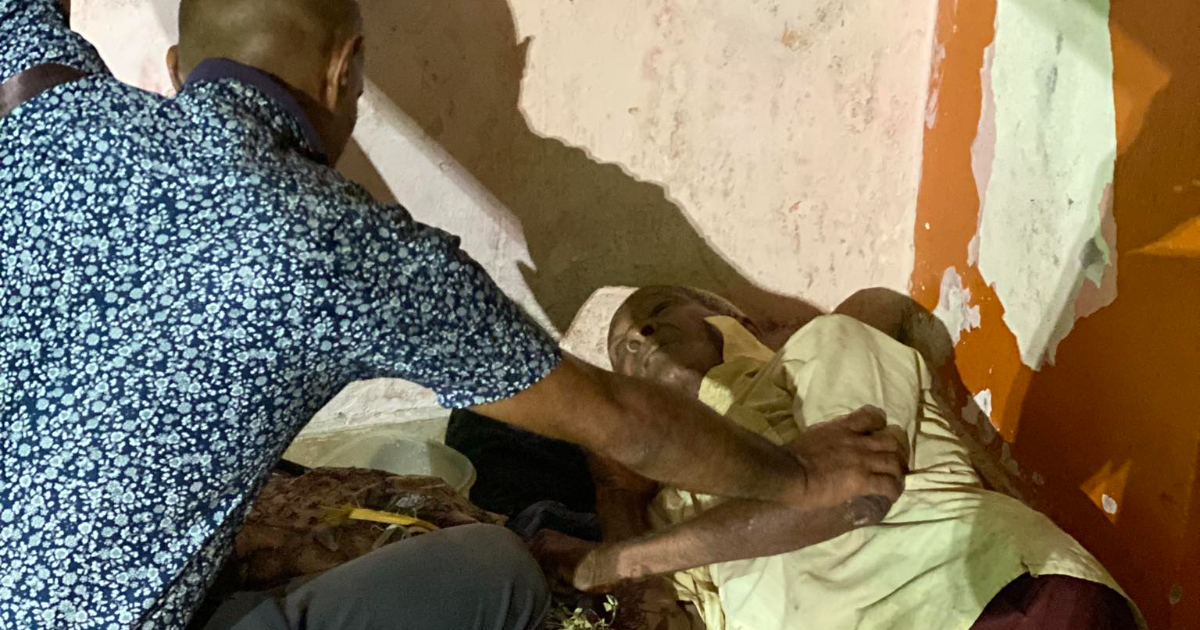 Fotografía conceptual y realista de Yasser Sosa Tamayo interactuando con un anciano cubano, el profesor Manuel, en un entorno urbano de Santiago de Cuba que evoca abandono y pobreza. Estilo cinematográfico, alta definición.