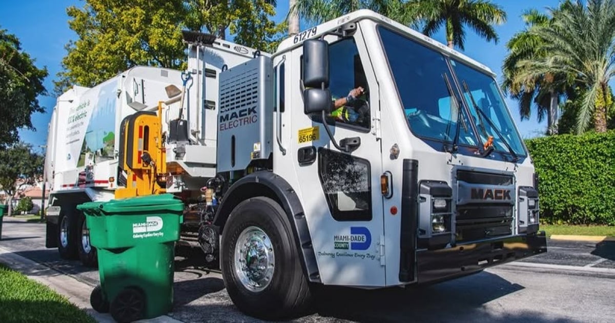 Fotografía fotorrealista de un moderno camión de recogida de basura del Condado de Miami-Dade, operando en una calle residencial soleada. El camión es azul oscuro con el logo del condado visible y un trabajador de saneamiento en el fondo. Estilo documental, alta definición.