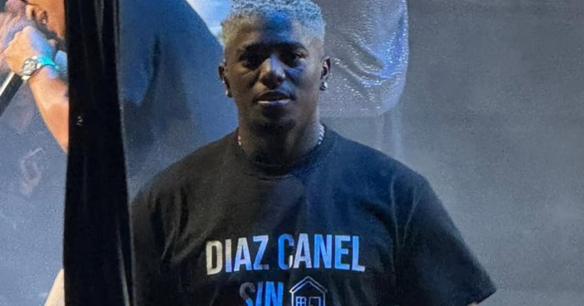 Fotografía fotorrealista de alta calidad de L Kimii en el escenario del Watsco Center en Miami, vistiendo una camiseta oscura con un mensaje político explícito contra Miguel Díaz-Canel. El artista está rodeado de luces de concierto y la multitud se ve al fondo, capturando el significado e impacto de la noticia. Estilo documental y realista.